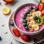 "A vibrant and delicious pitaya bowl topped with fresh fruits like sliced bananas, kiwi, and berries, garnished with granola and coconut flakes, served on a stylish table."